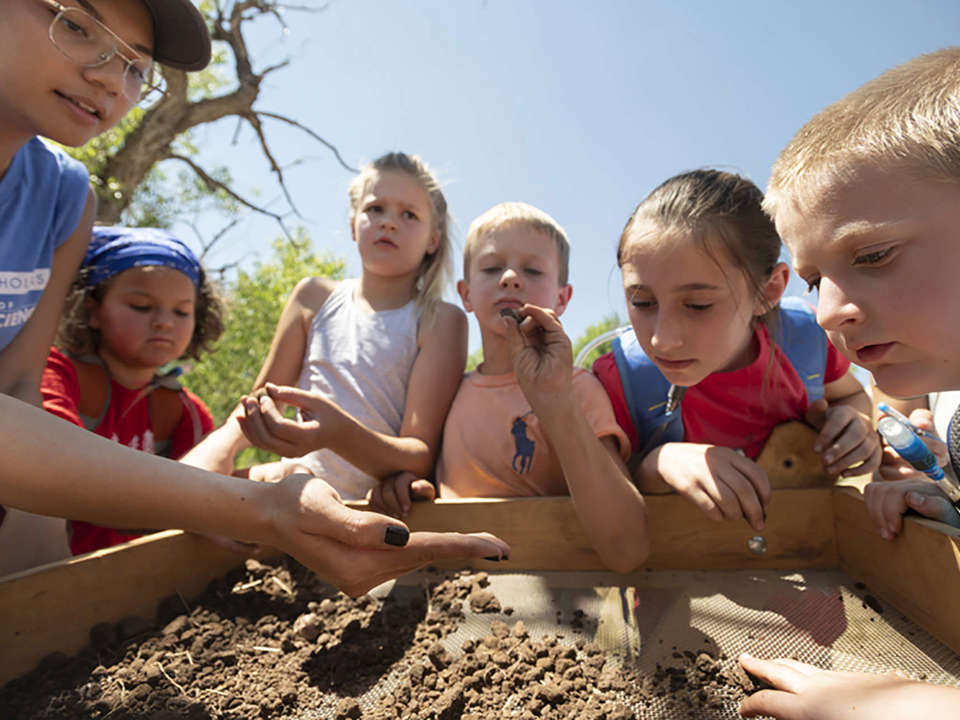 A group of five elementary school kids work with a volunteer to screen dirt to look for cultural materials