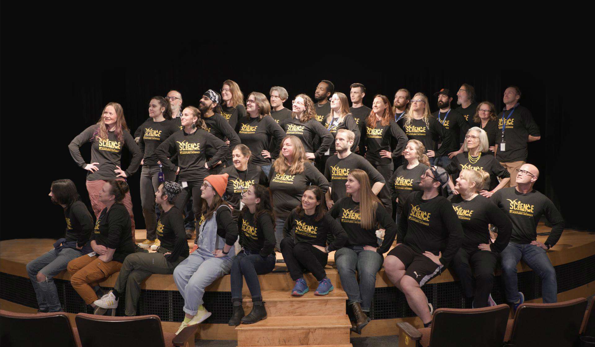 a large group of staff all wearing Science Division shirts stand on a stage with their hands on their hips looking off to the right.