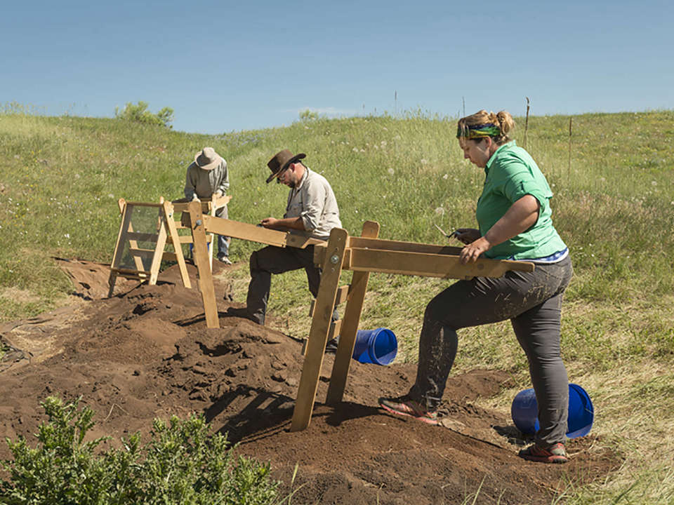 Three archaeologists work to screen dirt at the Magic Mountain site on a sunny day