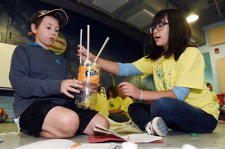 Two students seated on the floor use recyclable materials to create a model