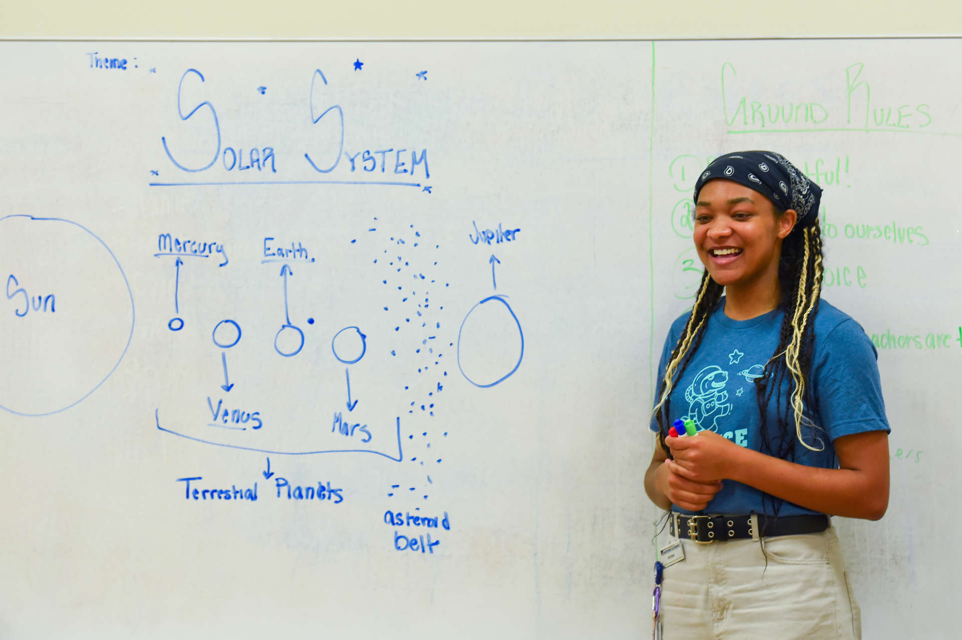 A teacher stands next to a white board with a partial drawing of the solar system.